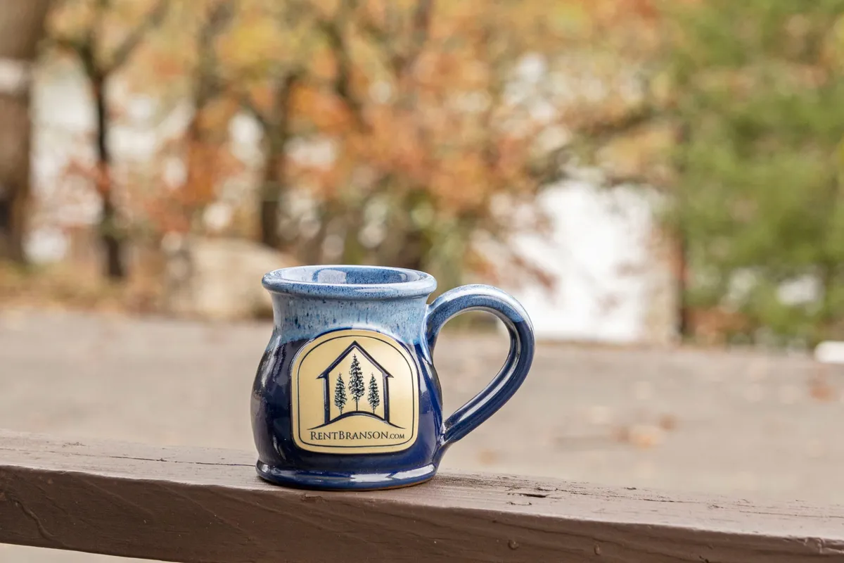 A close-up of a ceramic mug set against natural foliage, suggesting the experience of enjoying a coffee outside in a peaceful, green setting. The blurred background emphasizes tranquility and a cozy connection to nature.