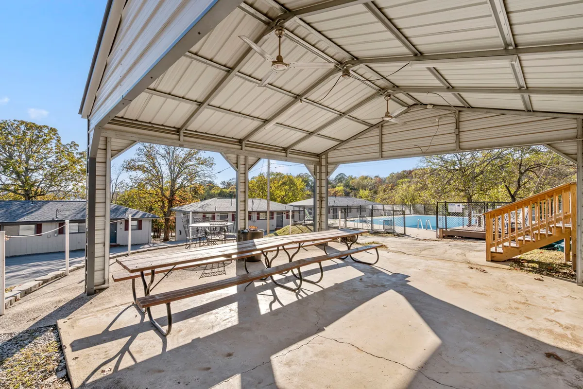 This exterior image showcases a covered patio with a picnic table, surrounded by trees and with a swimming pool visible in the background. The space looks inviting for both people and pets, combining shade, open air, and direct access to outdoor amenities.