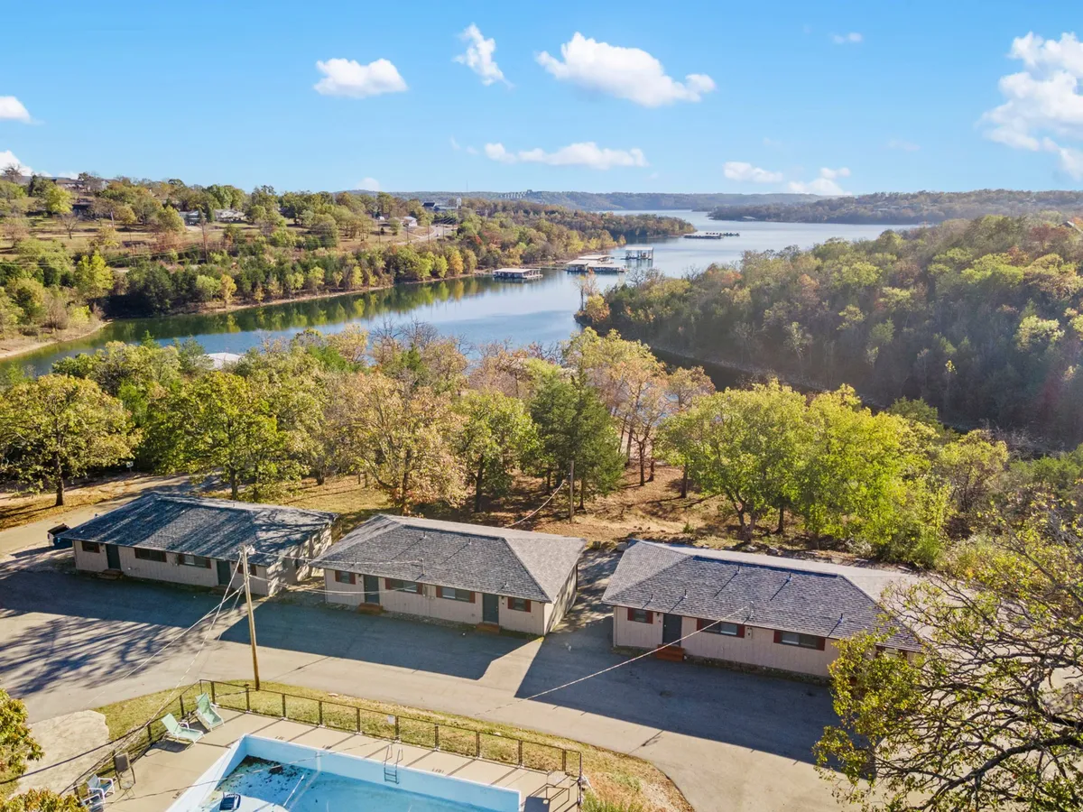 This exterior scene showcases a serene lakeside setting with a view of the swimming pool, cozy cabins, and Table Rock Lake in the background. The surrounding trees and open sky add to the tranquil, romantic atmosphere.