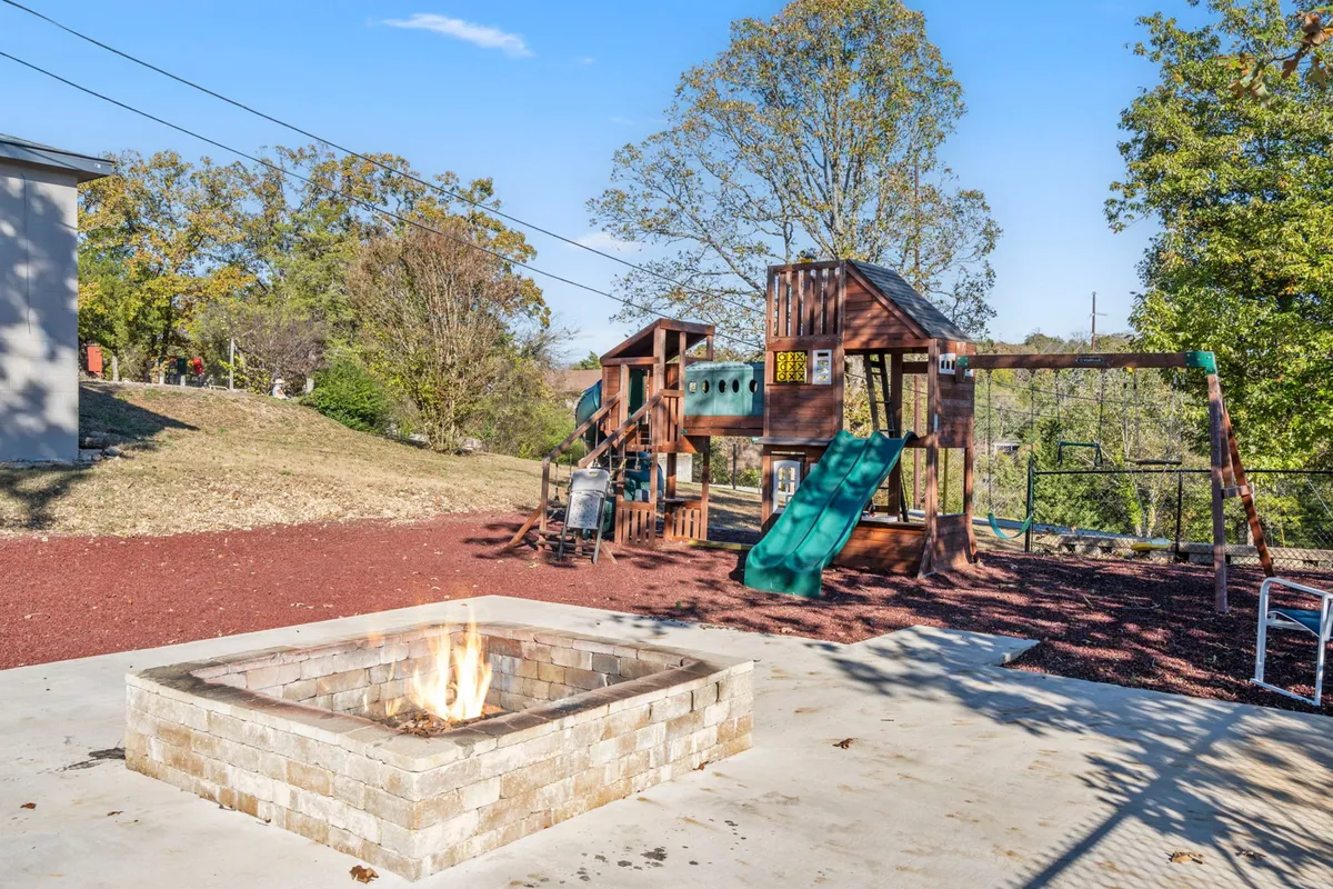 An exterior shot featuring a colorful playground structure and a fire pit in the foreground. The area is surrounded by greenery, inviting families to gather for playtime or evening marshmallow roasts by the fire.
