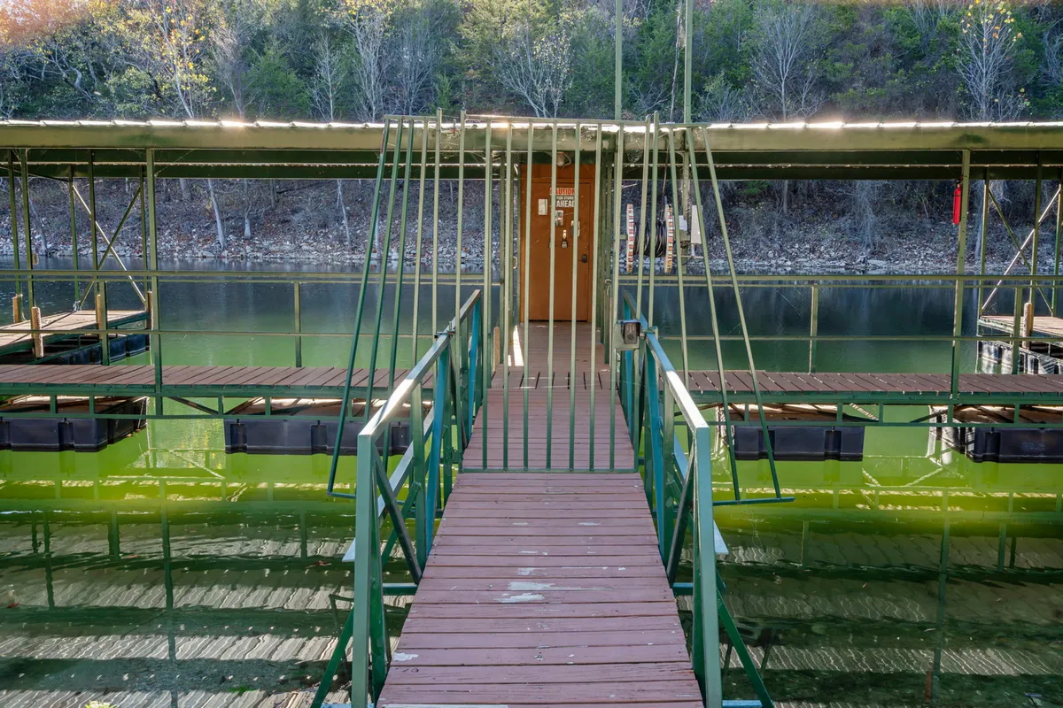 A tranquil wooden walkway leads to a dock above glistening water, surrounded by green railings and reflected light. The dock invites fishing, boating, and peaceful moments by the lakeside.