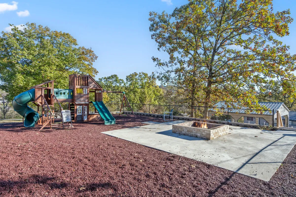 The image spotlights a vibrant playground structure adjacent to a fire pit, all shaded by large trees. The scene is alive with the potential for children's play, group gatherings, and cozy evenings around the fire.