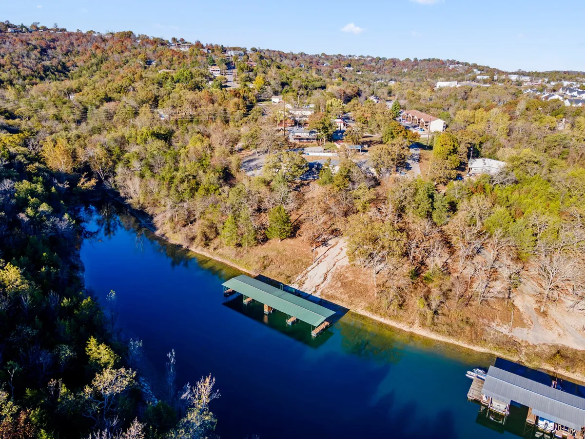 A wide view from the dock over the sparkling water, framed by trees, gently rolling hills, and a few houses in the background. The scene is peaceful, natural, and inviting, emphasizing the scenic beauty and recreational possibilities of the lake environment.