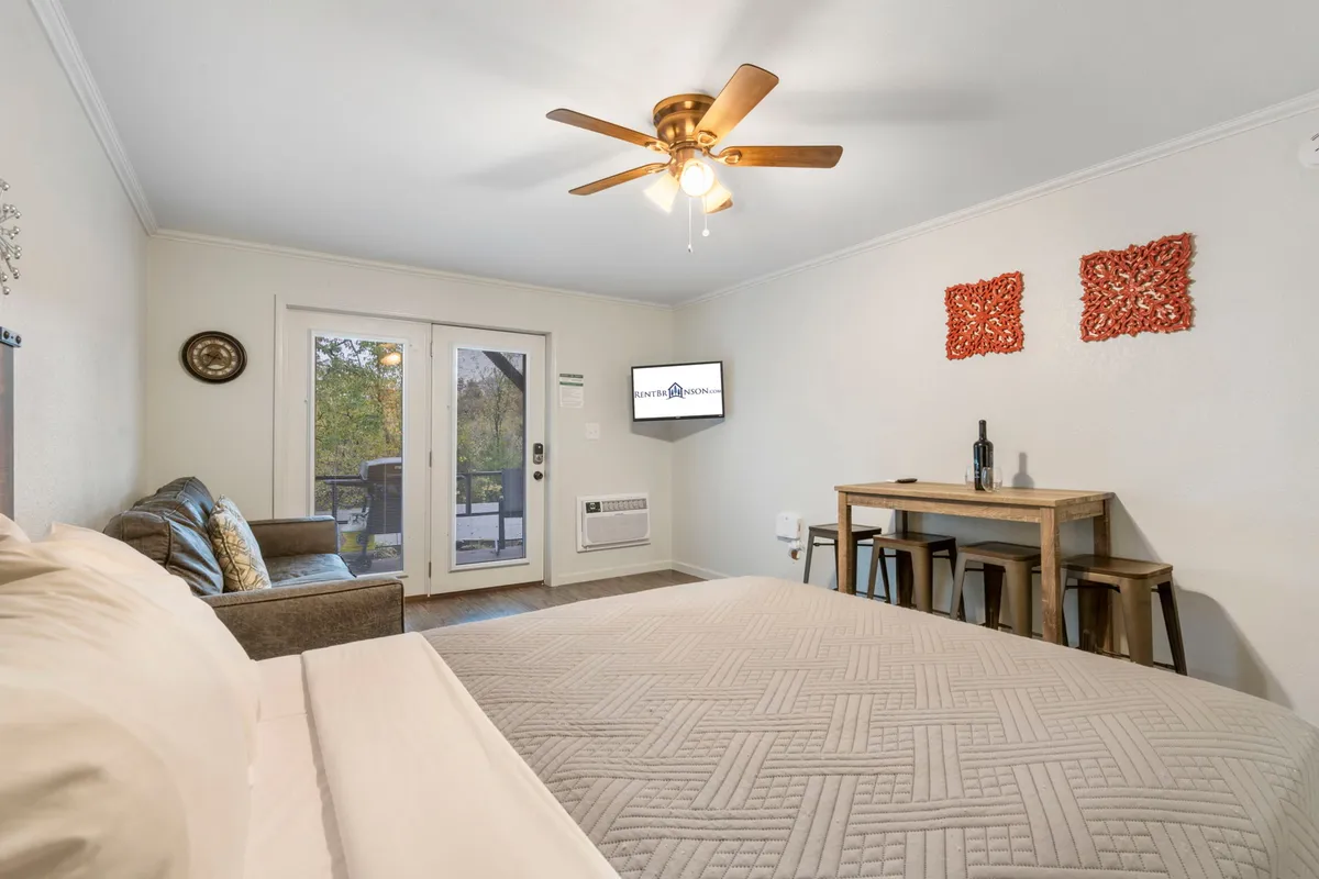 This inviting interior photo shows a plush queen bed, a cozy sofa, a small dining table, and sliding doors that lead to a patio. Decorative wall art adds a homely touch, while sunlight streams in, highlighting the spaciousness and warmth of the studio.
